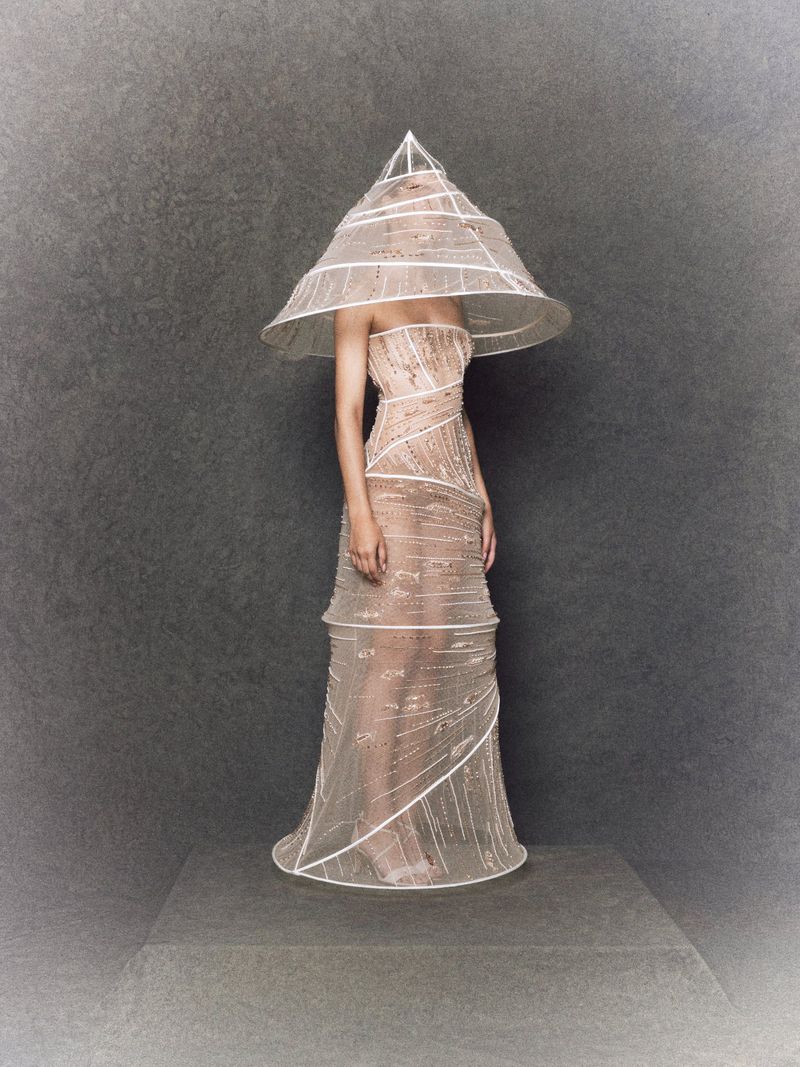 A person stands on a gray platform in a beige gown with a cone-shaped hat, embellished with white trim, against a mottled gray background.