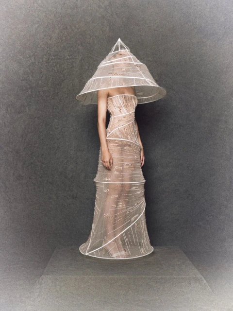 A person stands on a gray platform in a beige gown with a cone-shaped hat, embellished with white trim, against a mottled gray background.