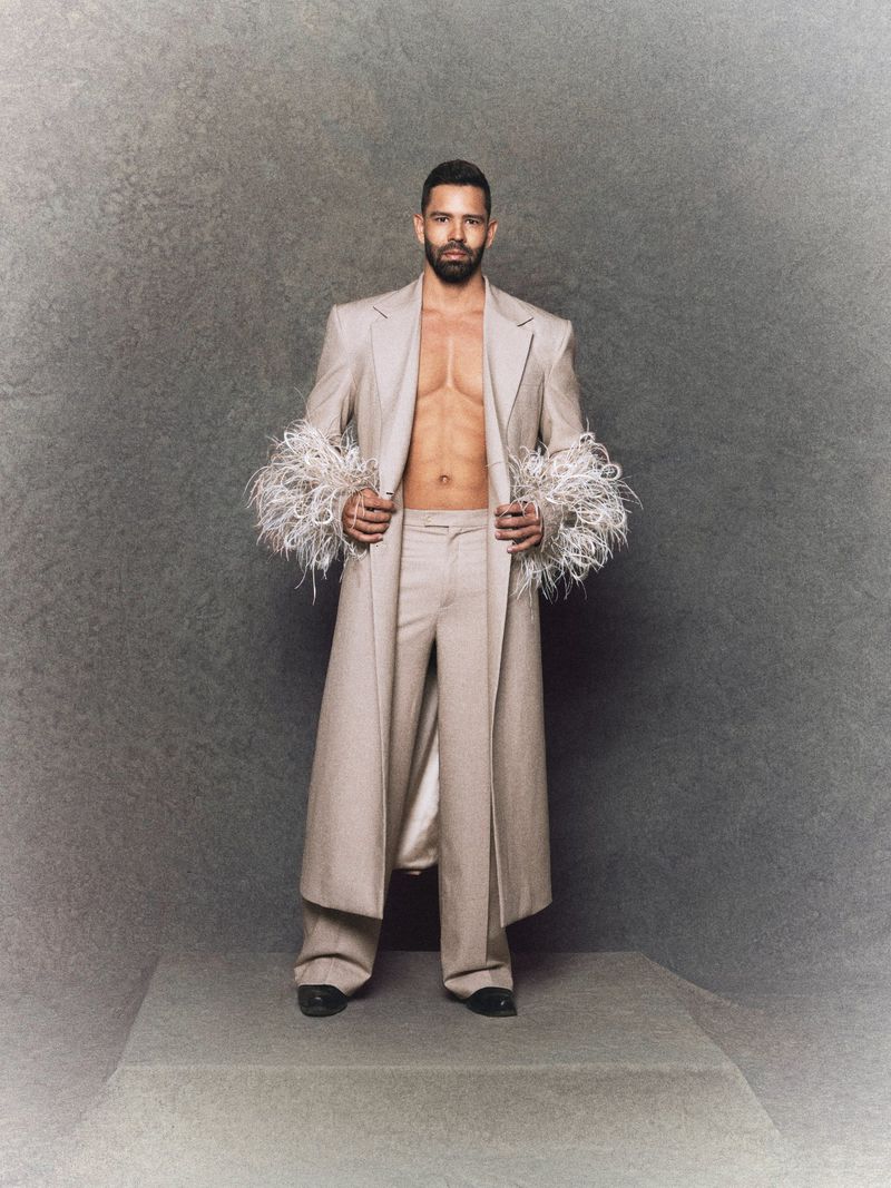 An adult man in a light-beige suit with feather trim poses in a studio setting against a mottled gray background, looking directly at the camera.