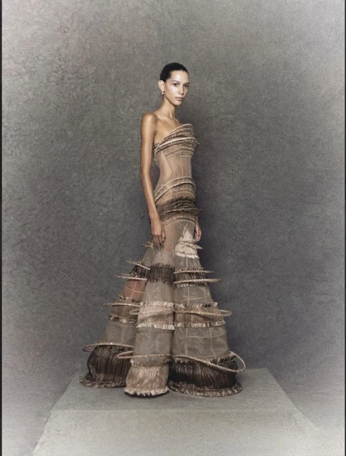 Elegant woman stands in a couture gown with sculptural ruffles and sheer fabric, posing against a textured grey backdrop in a studio setting.