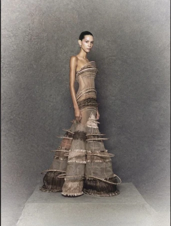 Elegant woman stands in a couture gown with sculptural ruffles and sheer fabric, posing against a textured grey backdrop in a studio setting.