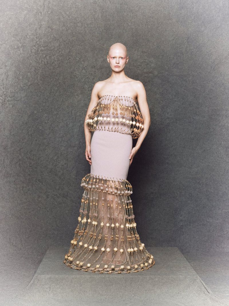 A bald adult female wearing a light pink couture gown with gold embellishments, standing in a full shot against a grey studio backdrop.