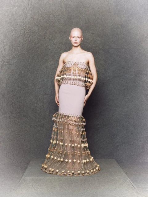 A bald adult female wearing a light pink couture gown with gold embellishments, standing in a full shot against a grey studio backdrop.