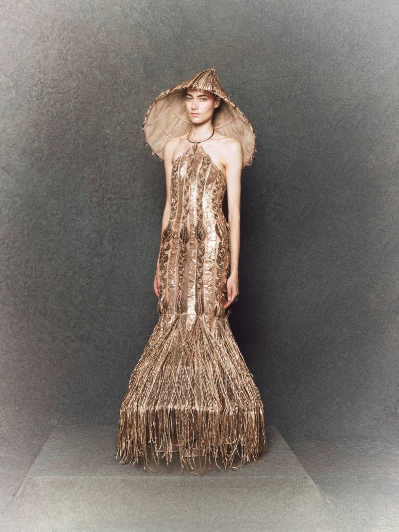 A fashion model presents a golden, fringed gown and wide-brimmed hat on a neutral gray stage, showcasing a blend of elegance and craftsmanship.
