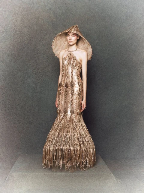 A fashion model presents a golden, fringed gown and wide-brimmed hat on a neutral gray stage, showcasing a blend of elegance and craftsmanship.