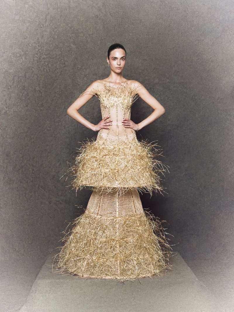 Fashion model poses wearing an elaborate couture gown of tiered straw ruffles against a mottled grey background in a studio fashion shot.