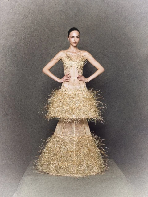 Fashion model poses wearing an elaborate couture gown of tiered straw ruffles against a mottled grey background in a studio fashion shot.