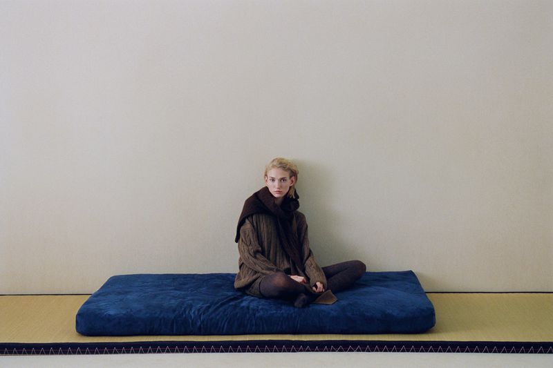 Woman with blonde hair sits cross-legged on a blue cushion against a neutral wall, wearing a brown sweater and scarf, indoors.