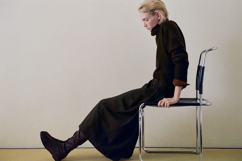 Model Tess Breeden wearing a dark brown turtleneck and matching skirt seated on a chrome chair against a neutral studio wall.