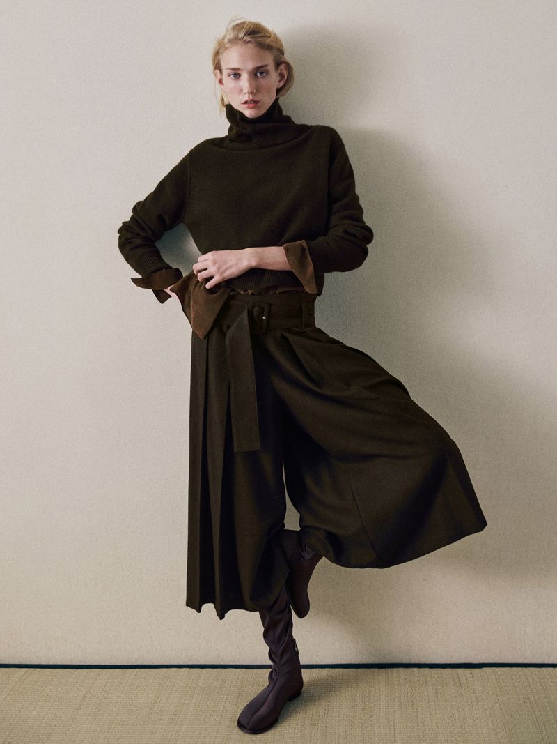 A stylish fair-skinned woman poses indoors, balancing on one leg. She wears an olive-green turtleneck, wide-leg pants with a belt, and matching boots.