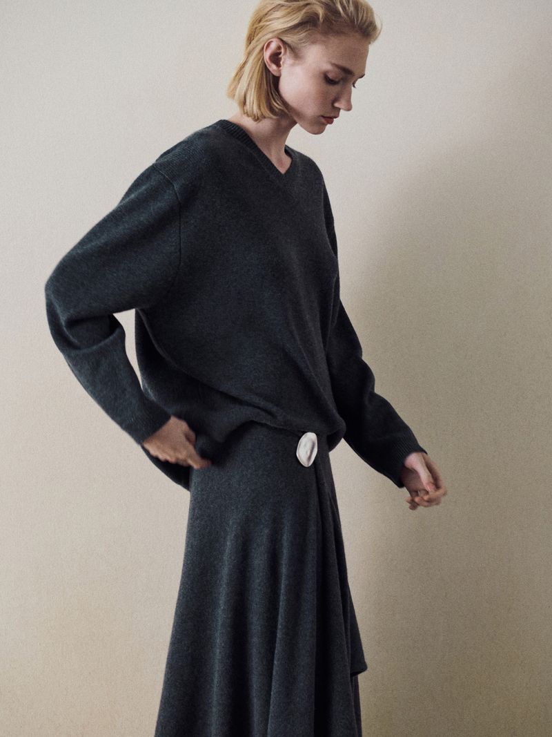 A slender woman models a dark grey V-neck sweater and a matching long, draped skirt against a neutral backdrop in a studio fashion shot.