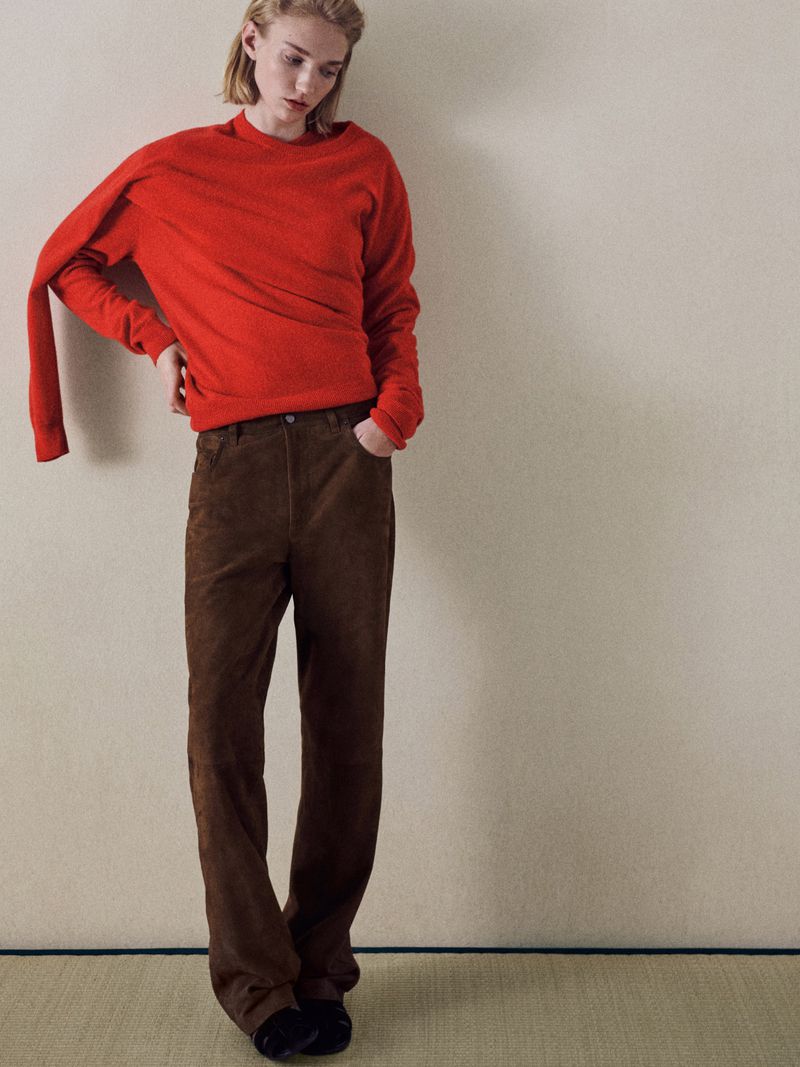 An adult posing indoors in a red sweater and brown corduroy pants, with one hand on her hip in a studio setting.