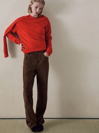 An adult posing indoors in a red sweater and brown corduroy pants, with one hand on her hip in a studio setting.