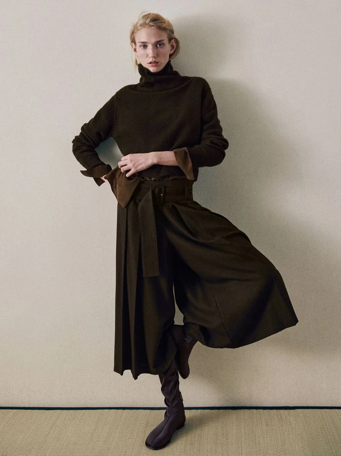 Model in dark turtleneck sweater and wide pleated trousers against a plain wall