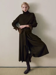 Model in dark turtleneck sweater and wide pleated trousers against a plain wall