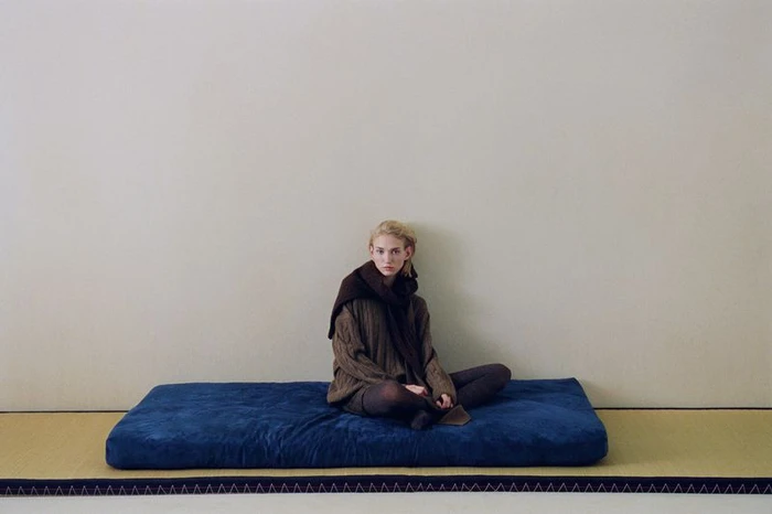 Tess Breeden seated cross-legged in a brown knit sweater and dark scarf on a blue cushion over a tatami mat against a neutral wall.