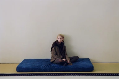 Tess Breeden seated cross-legged in a brown knit sweater and dark scarf on a blue cushion over a tatami mat against a neutral wall.