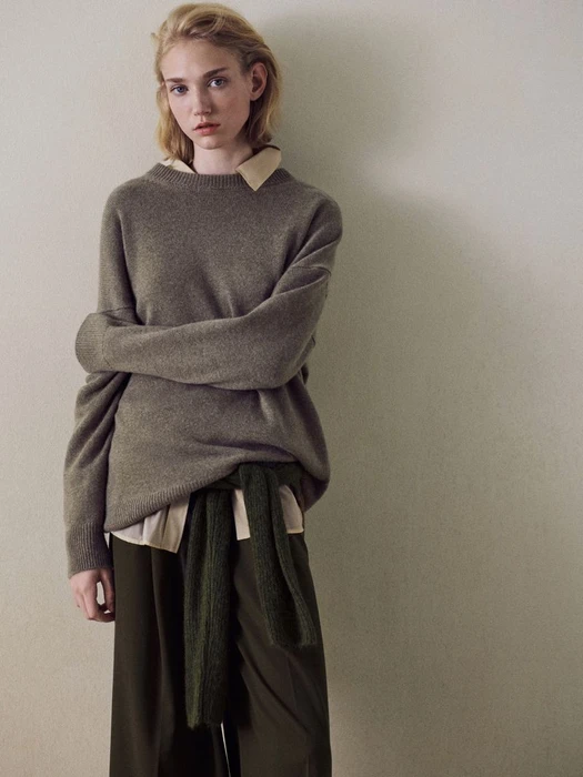 Model Tess Breeden posing in an oversized taupe cashmere sweater and dark olive trousers against a neutral studio backdrop.