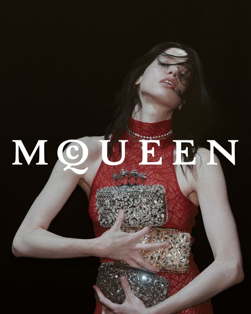 Woman in a red dress holds three embellished clutch purses against a black background in a studio fashion shot. Designer brand visible.