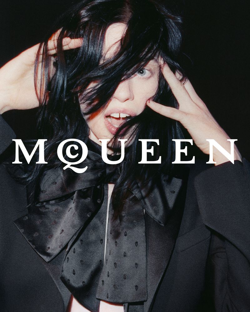 Fashion portrait of a woman with black hair wearing a black blazer with a bow tie, posing against a dark backdrop with the brand name MQEEN.