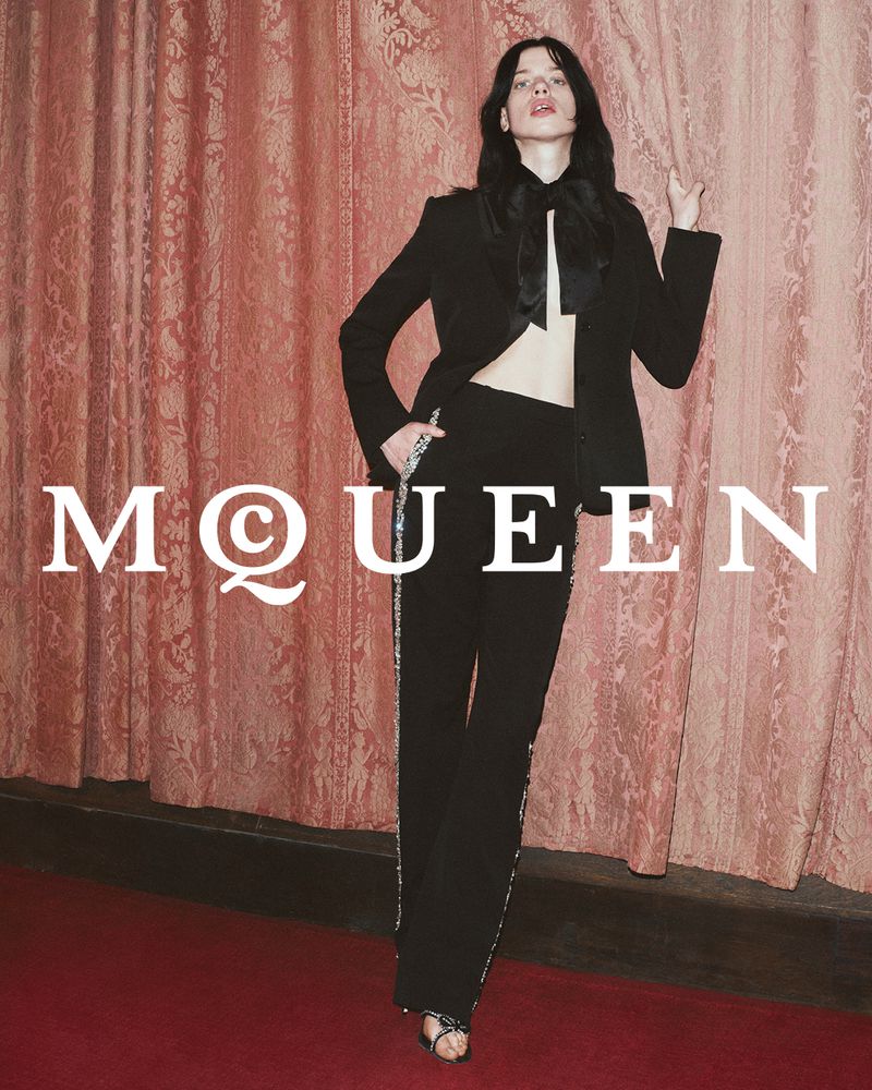 Model in black suit poses in front of coral damask curtain, full-length shot for Alexander McQueen fashion advertisement.