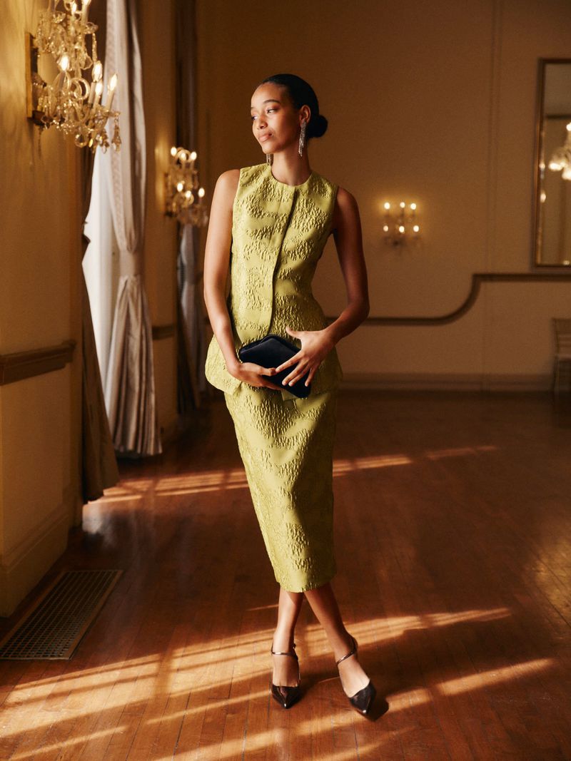 Woman in a textured chartreuse dress holding a black clutch, standing in a room with warm lighting and hardwood floors.