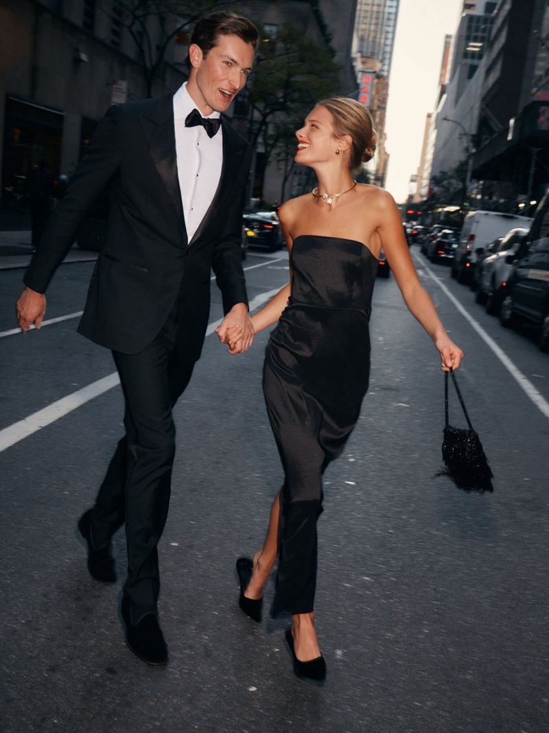 A smartly dressed couple in formal wear are walking on a city street, holding hands and smiling in an urban outdoor environment.
