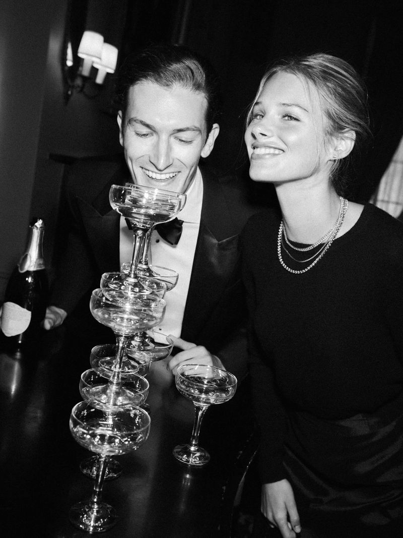A couple poses indoors by a tower of champagne coupes and a bottle in a black and white photograph with a dark, elegant atmosphere.