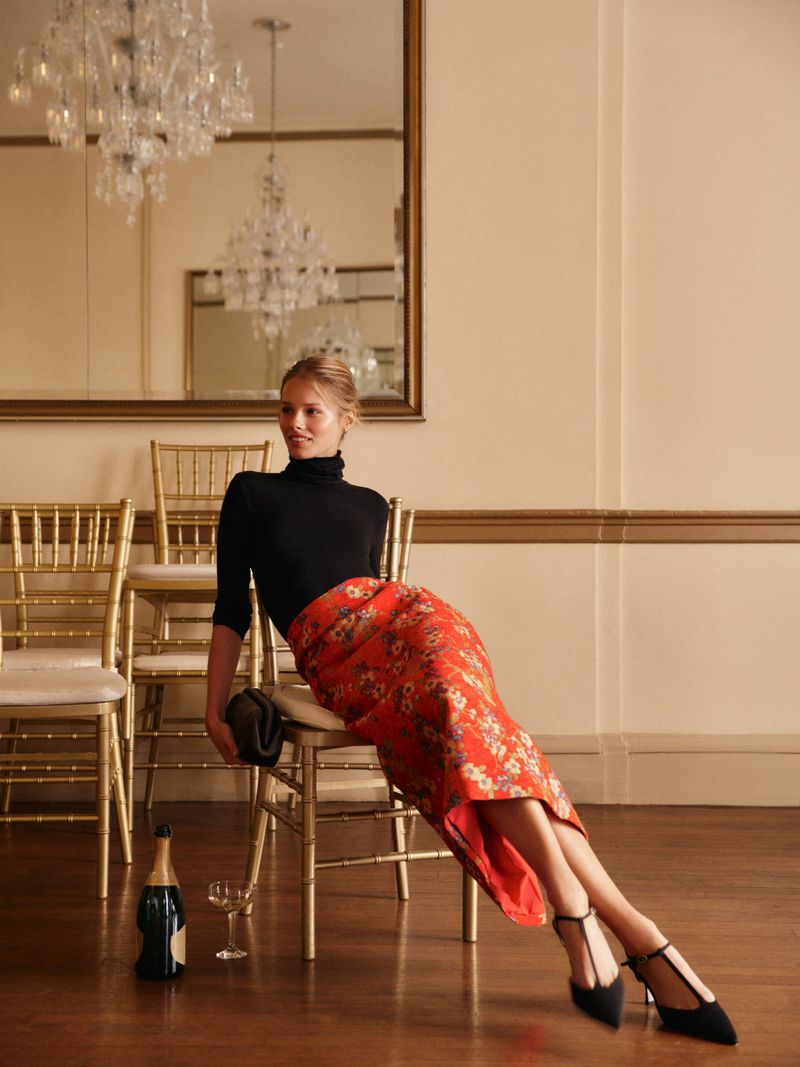 Woman in black top and red floral skirt sitting on gold chair in interior with chandeliers, champagne bottle, and wine glass.