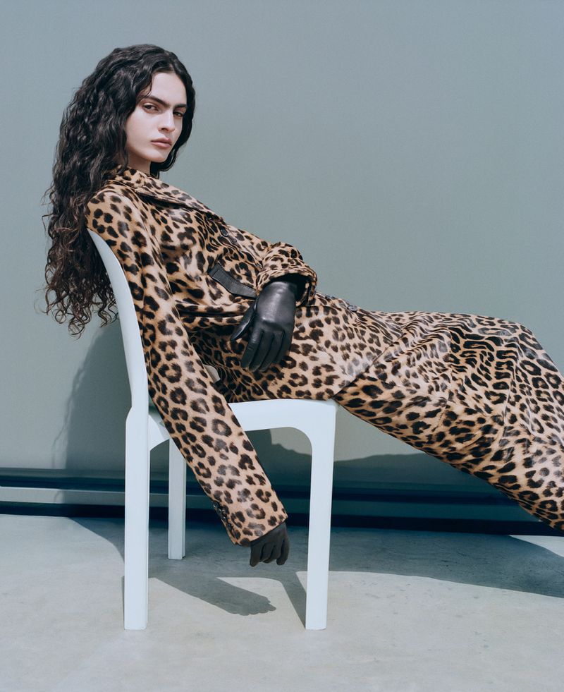 Woman with curly hair in a leopard-print suit poses on a white chair in a studio setting with a light blue background, wearing black gloves.
