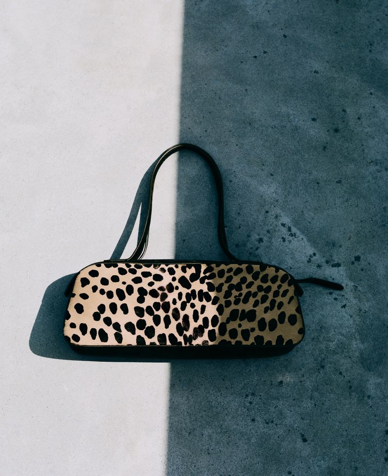 Overhead studio shot of a stylish handbag with black spots and a brown handle, casting a shadow on a split grey and white background.