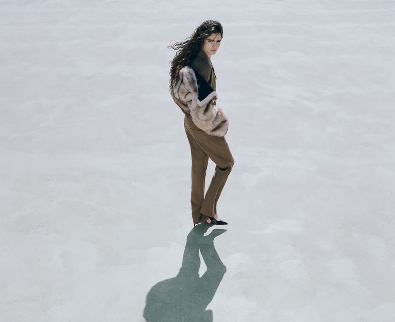Elegant woman in stylish tan suit and faux fur stole posing on a neutral surface with sunlight and shadow. Fashion editorial photography.