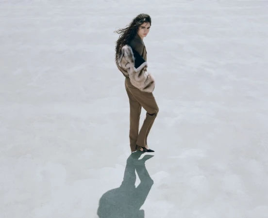 Elegant woman in stylish tan suit and faux fur stole posing on a neutral surface with sunlight and shadow. Fashion editorial photography.