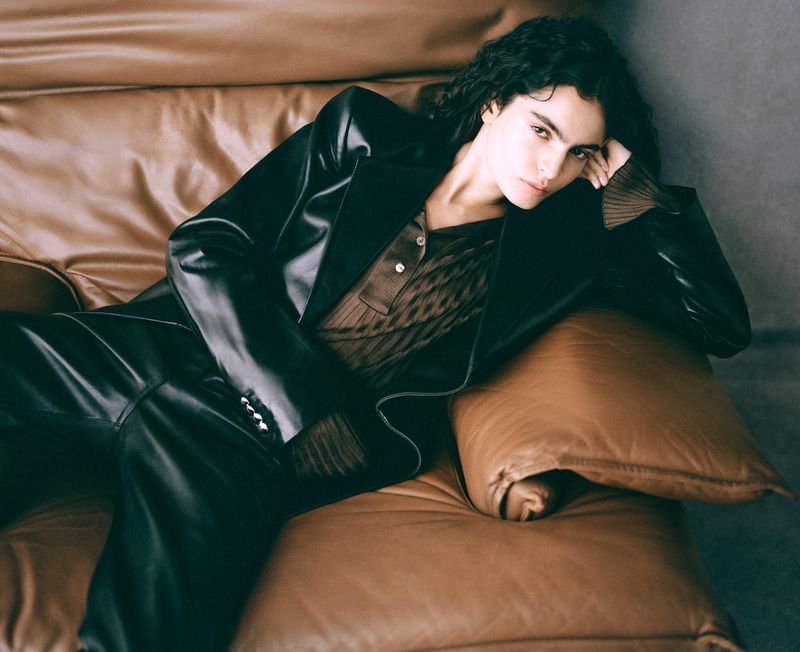 A brunette woman with curly hair lounges on a brown leather couch, wearing a dark blazer and brown top, looking at the camera.