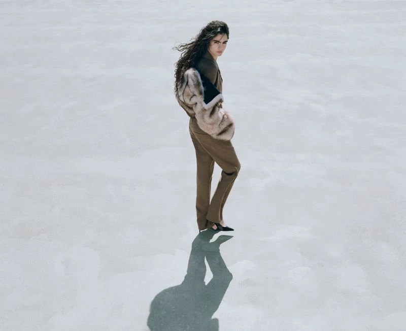 Model Carol Monteiro looking over her shoulder wearing a brown tailored suit and fur jacket on a pale minimalist floor