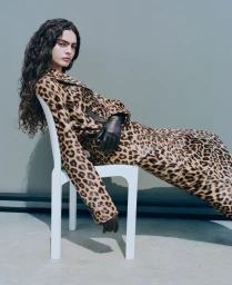 Model Carol Monteiro wearing a leopard print coat and black leather gloves while reclining on a white chair against a plain studio wall.