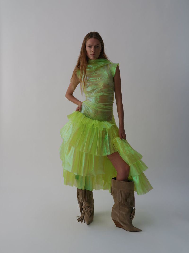 A woman poses in a translucent green high-fashion dress and tan fringe boots against a stark white backdrop in a fashion photo shoot.