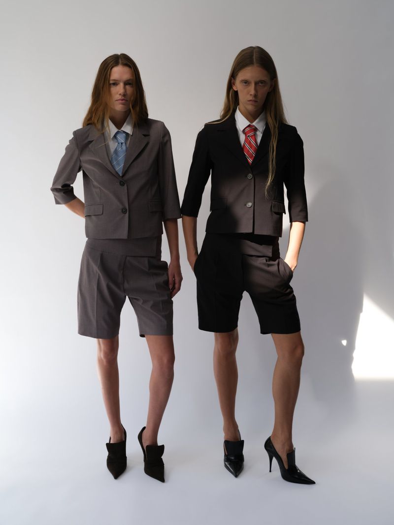 Two models pose in cropped blazers, shorts, and heels against a white backdrop in a studio, showcasing minimalist, business-casual fashion.