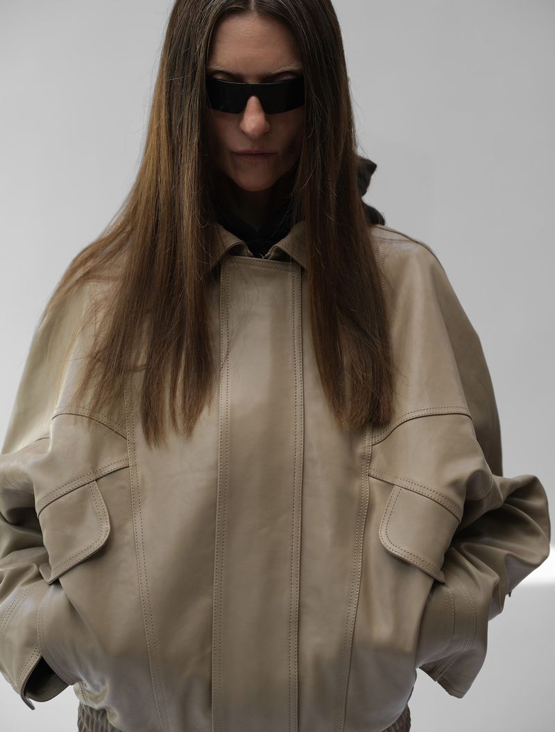A woman with long brown hair wears black sunglasses and a tan leather jacket against a plain white background in a studio shot.