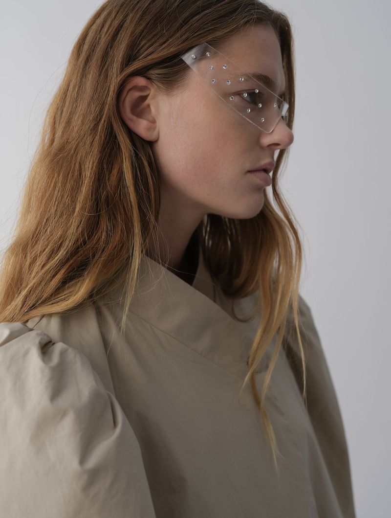 Profile portrait of a light-skinned woman with long auburn hair wearing transparent rhinestone-studded visor against a white background.