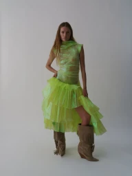 Model in neon green ruffled dress wearing fringed tan suede boots against a plain grey studio backdrop
