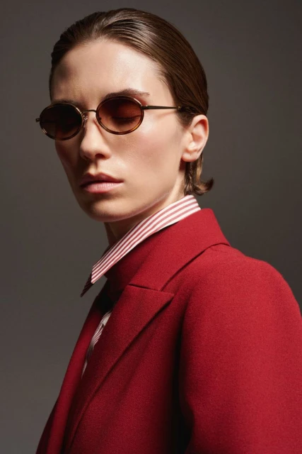 Stylish woman with short brown hair and red blazer wearing round, brown-tinted glasses against a grey studio backdrop.