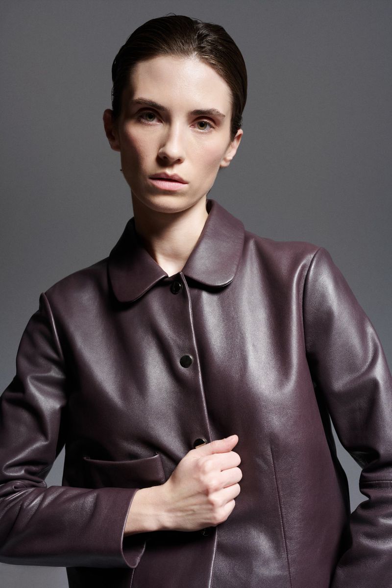 Model Cleo Cwiek wearing a deep burgundy leather jacket with a rounded collar against a neutral grey studio backdrop.