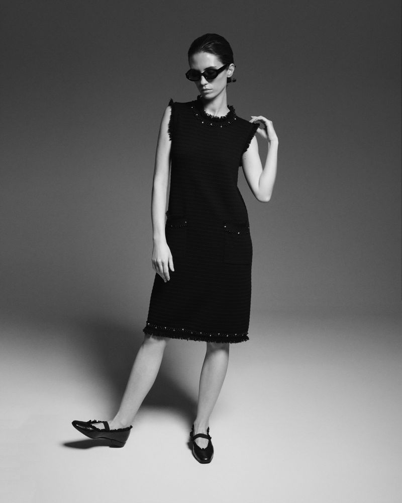 Black and white shot of a young adult woman posing in a black dress with sunglasses and black shoes in a photo studio.
