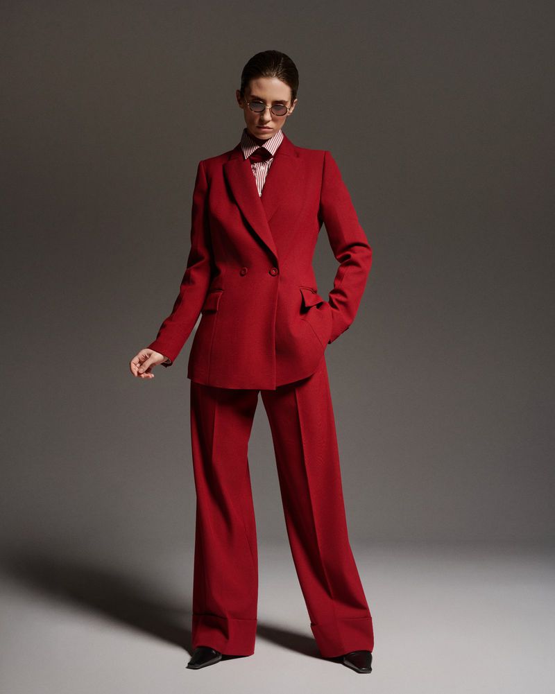 Model wearing a tailored red pantsuit and tinted glasses standing against a grey studio backdrop.