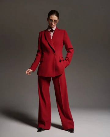 Model wearing a tailored red pantsuit and tinted glasses standing against a grey studio backdrop.