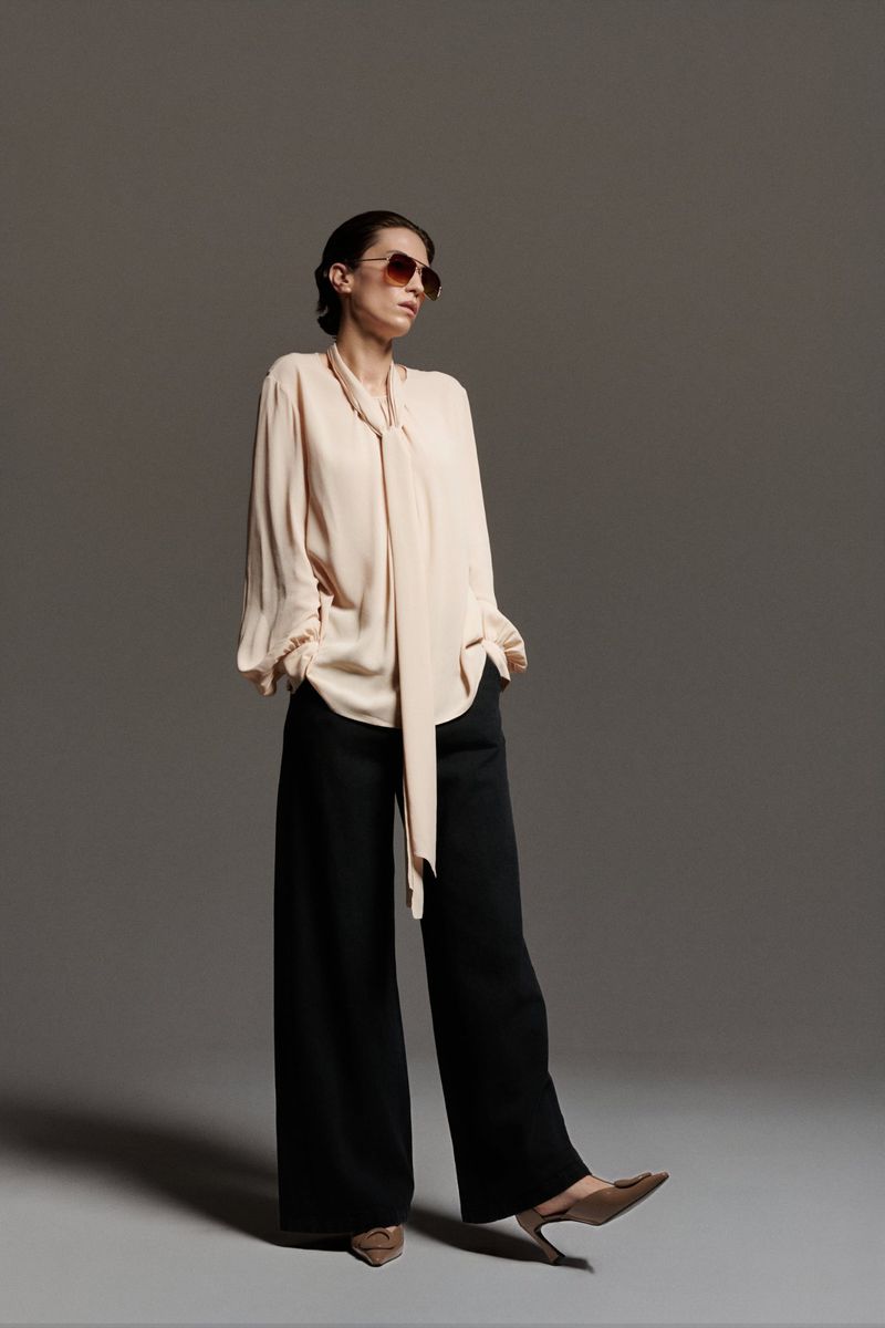 A woman poses in a peach blouse with wide-legged black pants and brown heels in a studio environment with a grey backdrop.