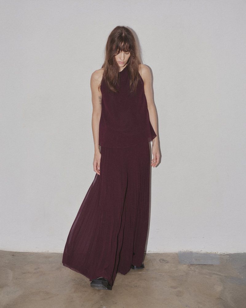 A woman with long brown hair models maroon palazzo pants and a sleeveless halter top in a minimalist indoor setting against a white wall.