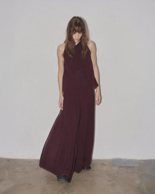A woman with long brown hair models maroon palazzo pants and a sleeveless halter top in a minimalist indoor setting against a white wall.