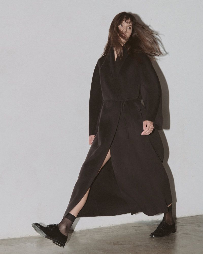 Woman in a long dark coat walks against a white wall, showcasing fashion and style with her dark dress and patent leather shoes.